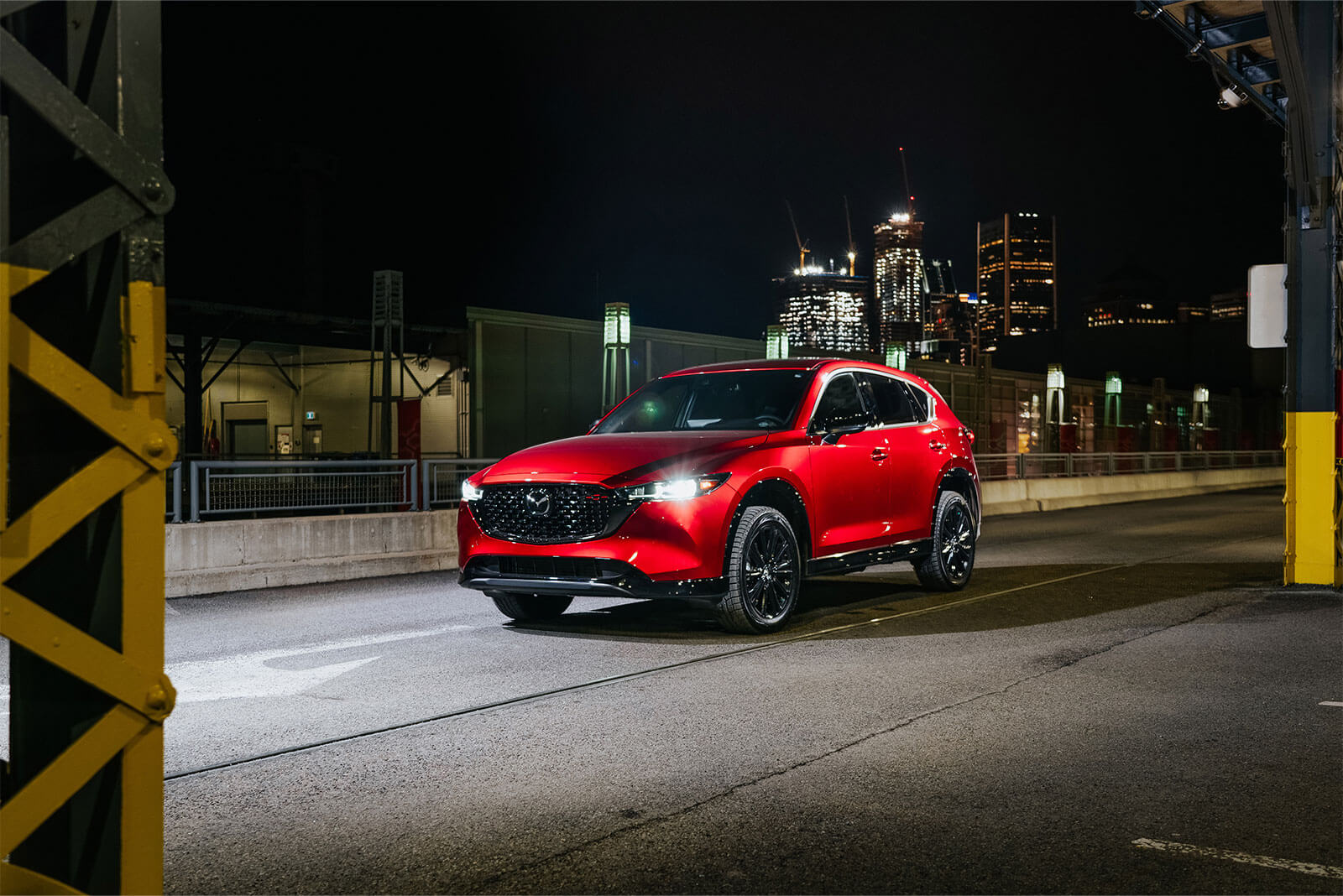 Soul Red Crystal Metallic Mazda CX-5 drives past blurred structures along urban street.