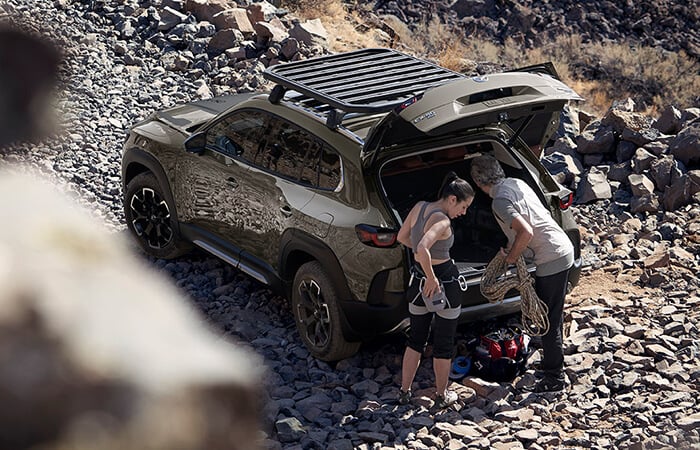 Birds-eye view of couple unloading outdoor gear from back of CX-50 parked on gravel track.