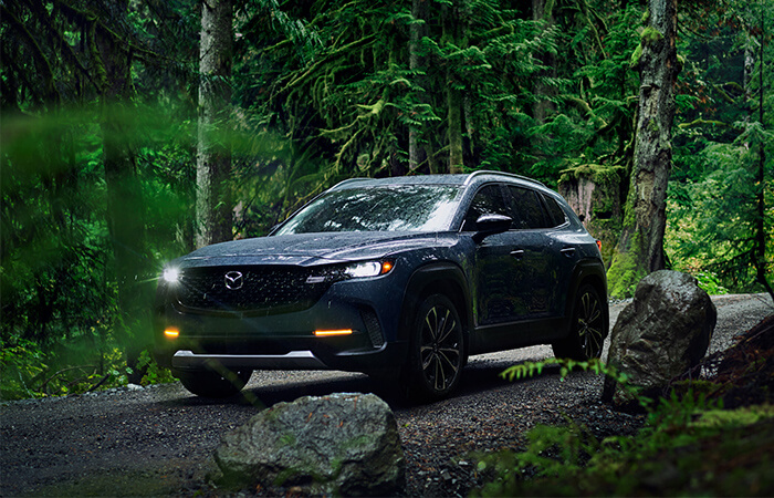 Ingot Blue Mica CX-50 with headlights on driving in dense forest.