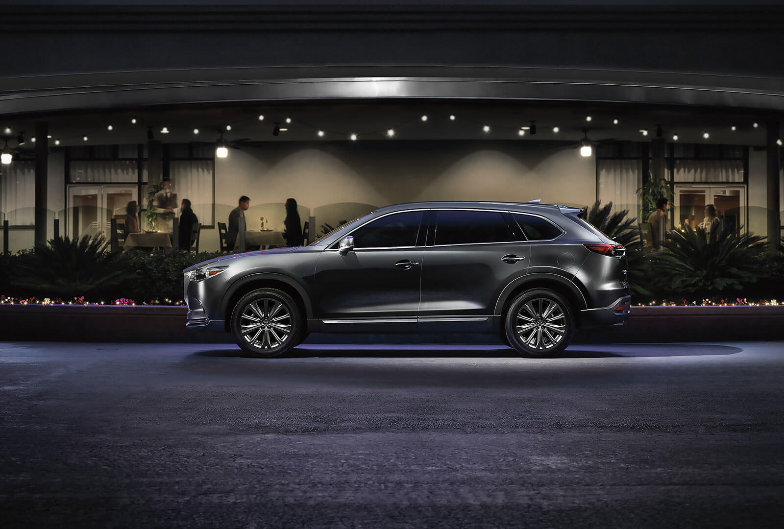 Polymetal Grey Metallic CX-9 reflects streetlights in front of a restaurant patio at night.