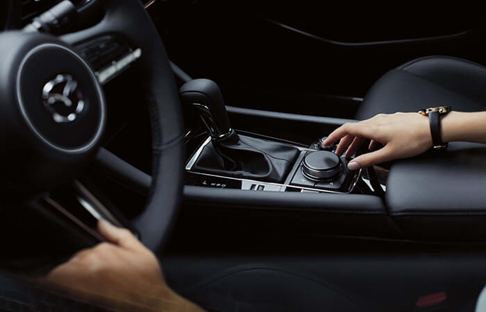 Driver in the Mazda3 cockpit, one hand reaches for the dial in the centre console while the other rests on the steering wheel.