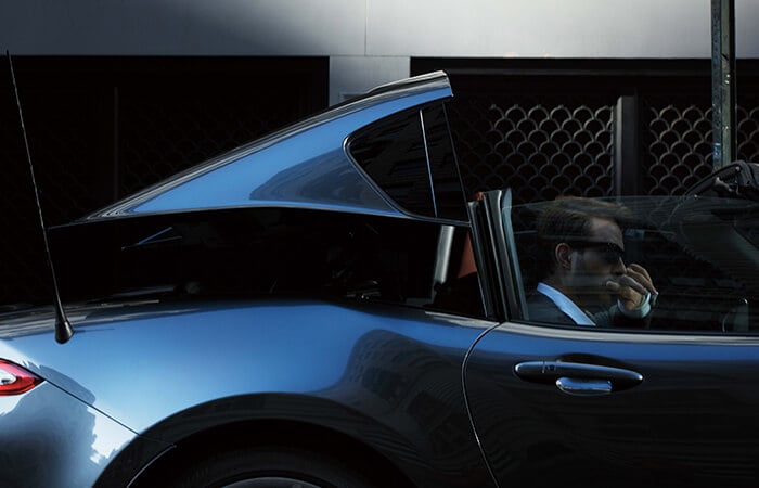 Un conducteur avec des lunettes de soleil baisse le toit de sa MX-5 RF gris mécanique métallisé garée.