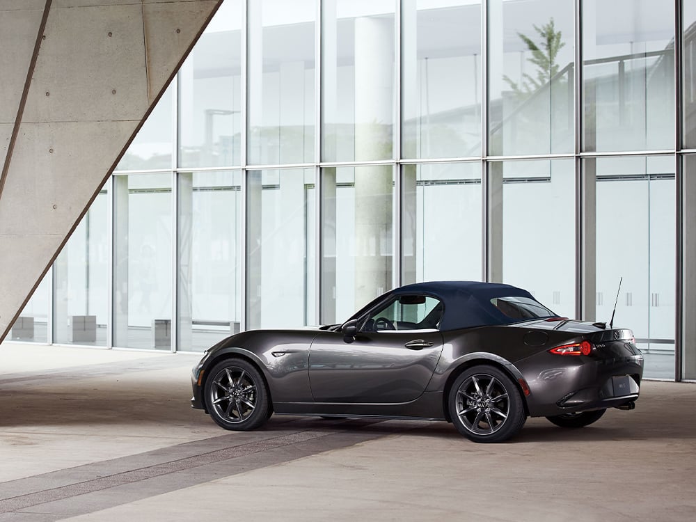 Machine Grey Metallic MX-5 Soft Top parked between bright concrete structure and tall glass building atrium.