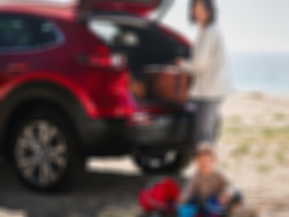 A woman pulls a picnic basket out of the rear of their CX-30 parked at the beach, a child plays with buckets in the sand. 