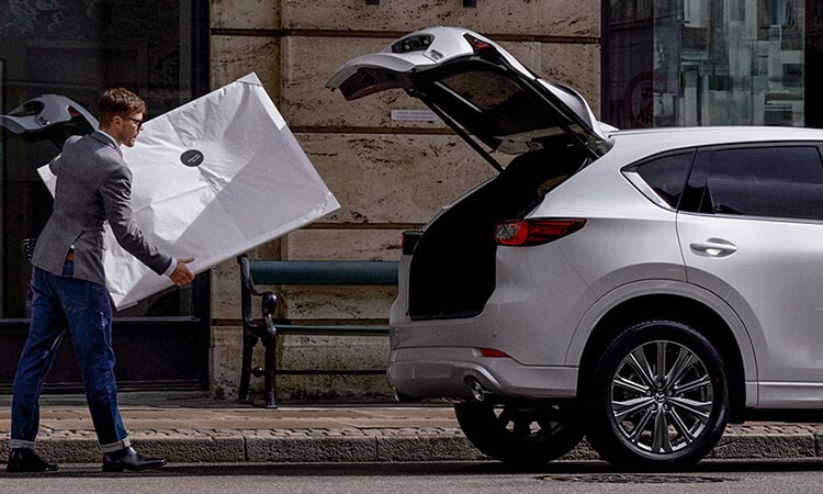 Un homme transporte un cadre blanc vers un Mazda CX-5 blanc rhodium métallisé garé dans la ville avec le hayon ouvert. 