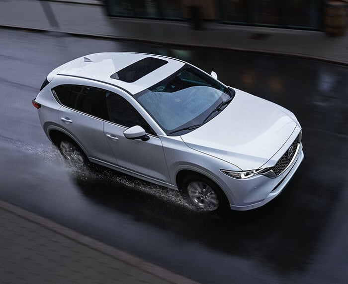 Un Mazda CX-5 blanc rhodium métallisé  s’éloigne sur un viaduc, devant des bâtiments de couleur marron. 