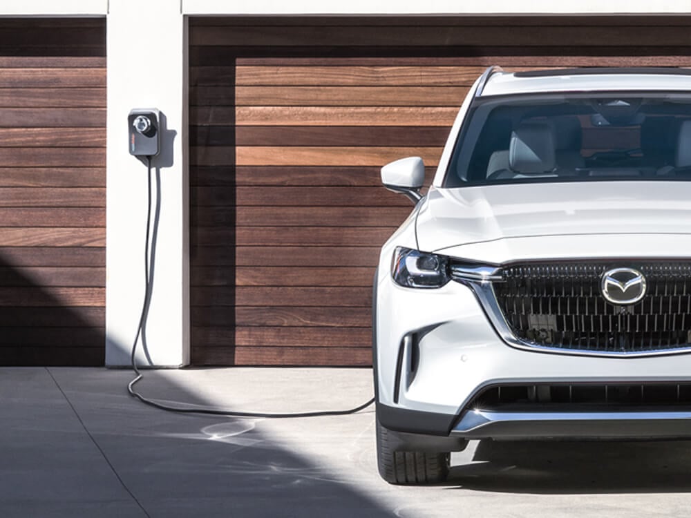 Plugged in and charging Arctic White CX-90 PHEV parked in front of double garage with wood slat garage doors. 