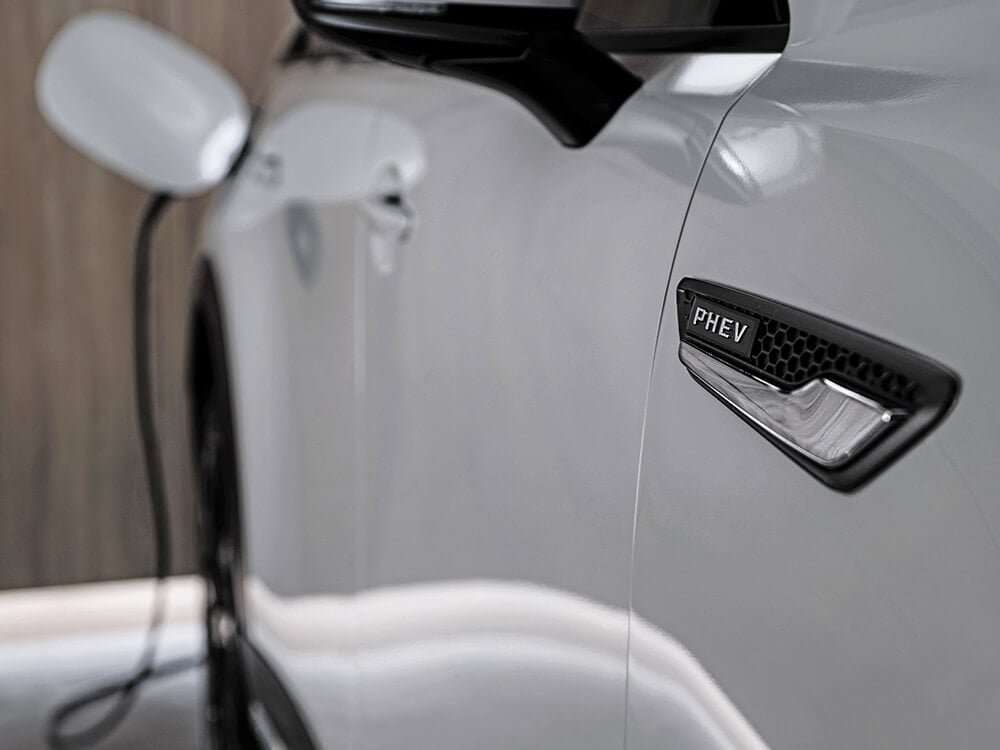 Close up of PHEV vehicle badge on the passenger side of Arctic White CX-90, charger plugged.  