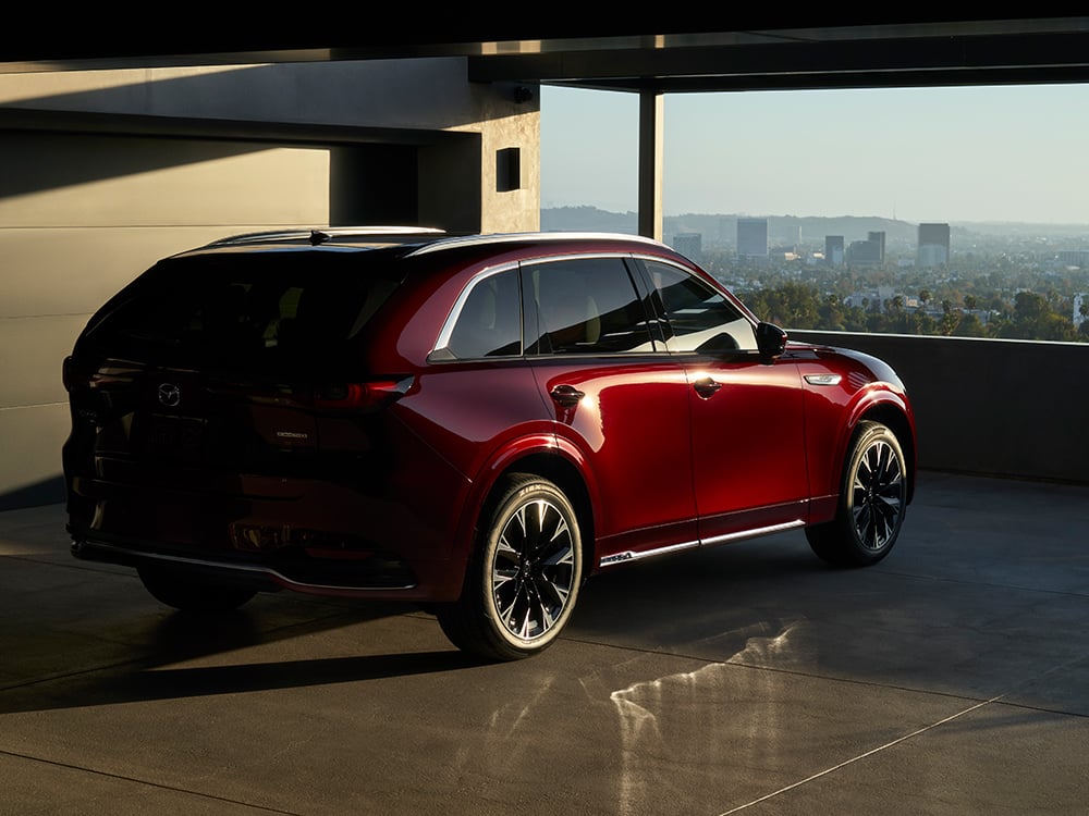 Un CX-90 rouge artisan garé dans un stationnement avec vue sur un paysage urbain au coucher du soleil.