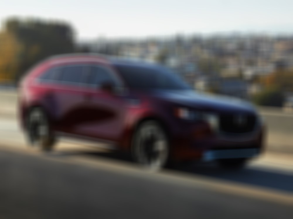 Artisan Red CX-90 traveling down concrete highway overpass reflecting daytime sunlight. 
