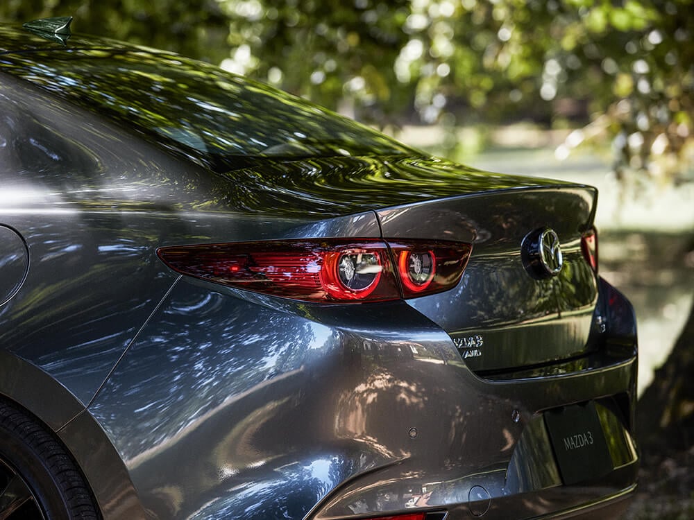 Une berline Mazda3 garée dans un parc verdoyant avec les feux arrière en gros plan lors d’une journée d’été ensoleillée. 