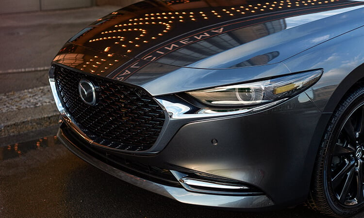 Phare du côté conducteur et capot d’une Mazda3 gris mécanique métallisé reflétant la lumière et les gratte-ciel dans une rue urbaine. 