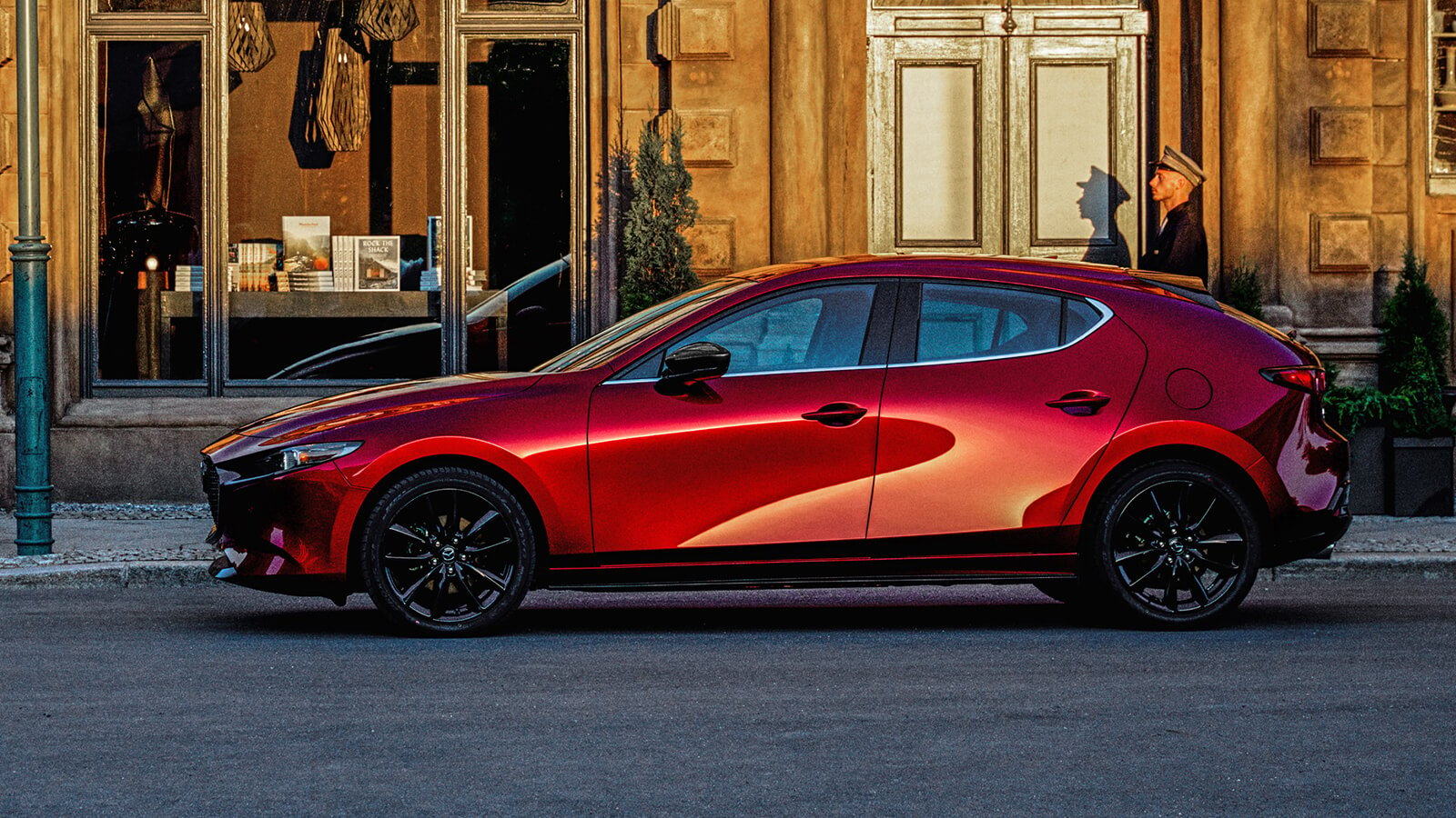 Une Mazda3 Sport rouge vibrant cristal métallisé garée devant un magasin de la rue principale au coucher du soleil.