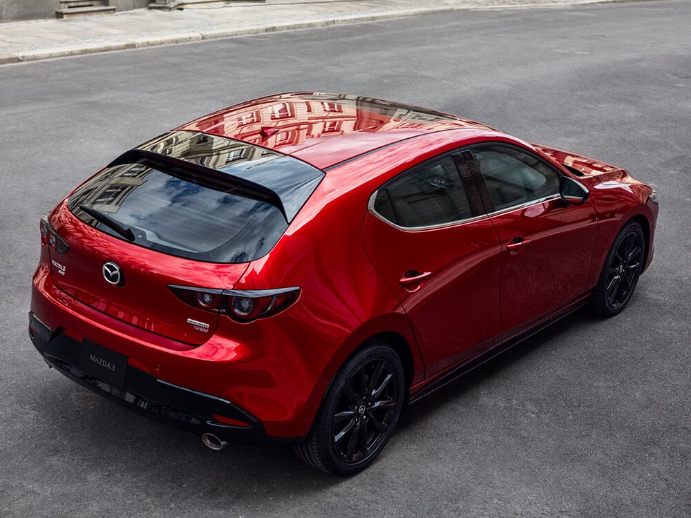 Vue en contre-plongée de l’arrière, côté passager, d’une Mazda3 Sport dont la couleur rouge vibrant cristal métallisé reflète les bâtiments environnants. 