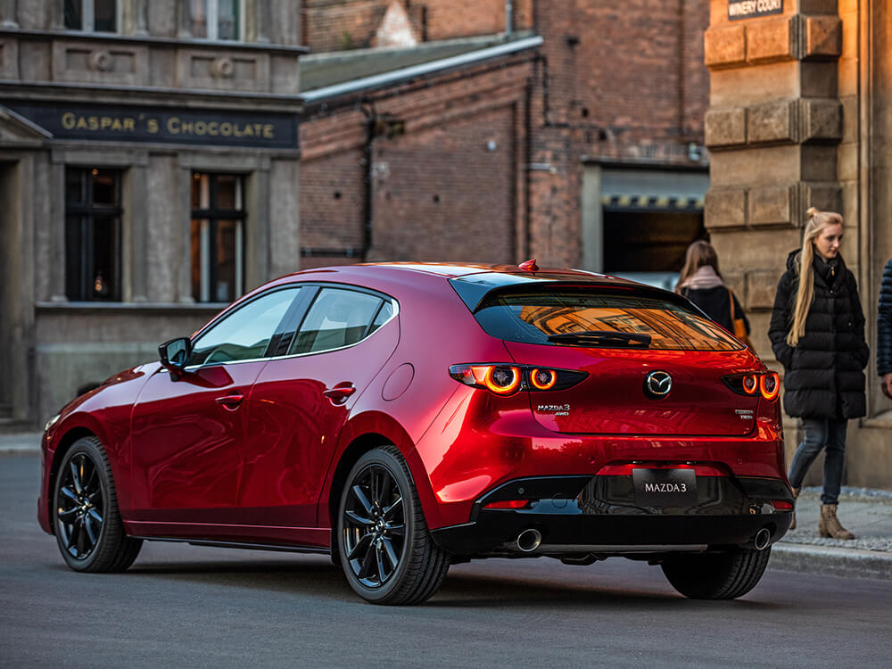 Une Mazda3 Sport rouge vibrant cristal métallisé arrêtée au coin de la rue principale d’une ville pittoresque; un couple passe sur le trottoir. 