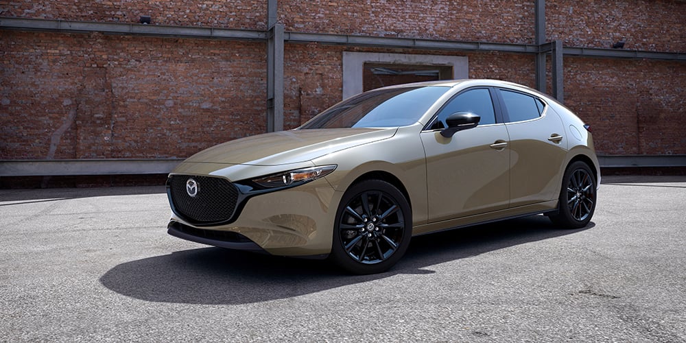 Zircon Sand Metallic Mazda3 Sport parked behind red brick warehouse, reflecting a clear sky in midday sunlight.