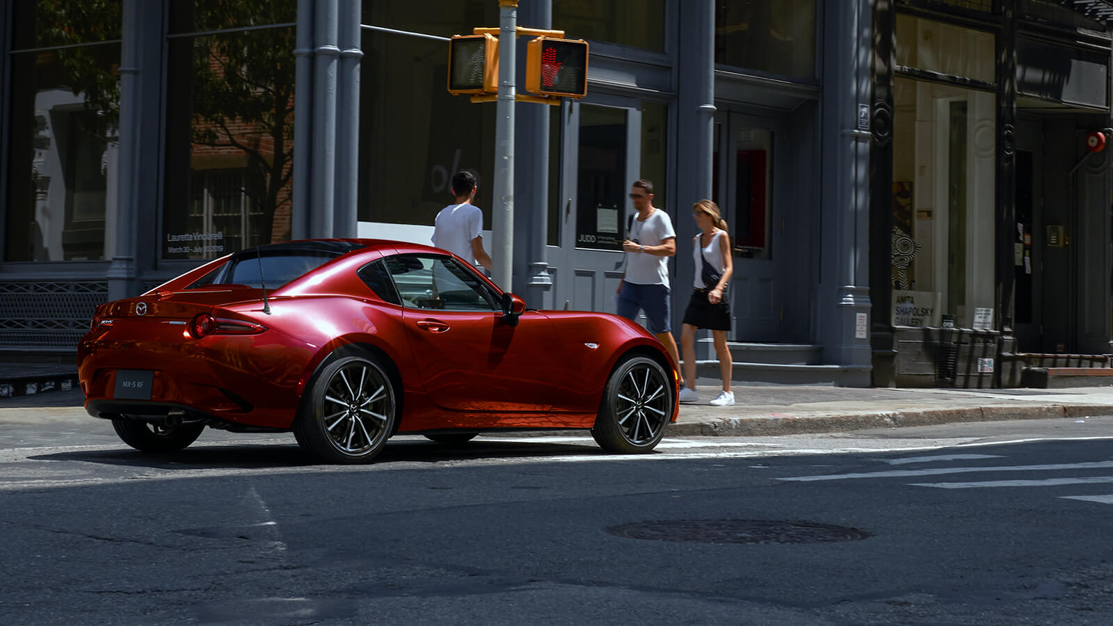 Soul Red Crystal Metallic MX-5 RF rests at a stop light on city main street, pedestrians walk by in the background.