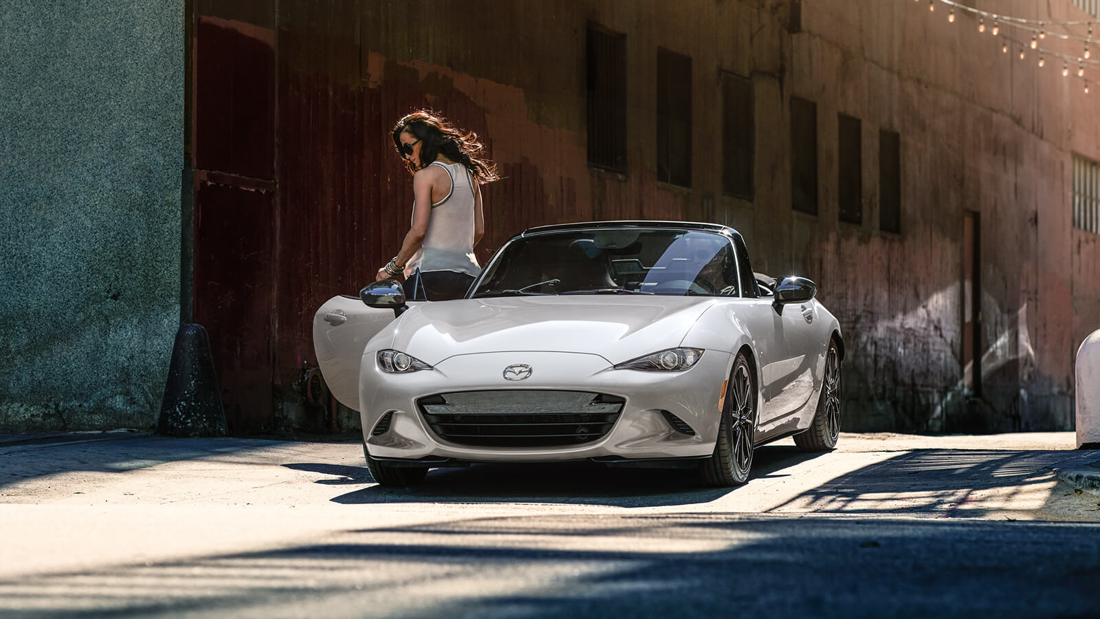 Woman exits the passenger's side of Snowflake Whit Pearl MX-5 parked in front of large, weathered wooden door. 