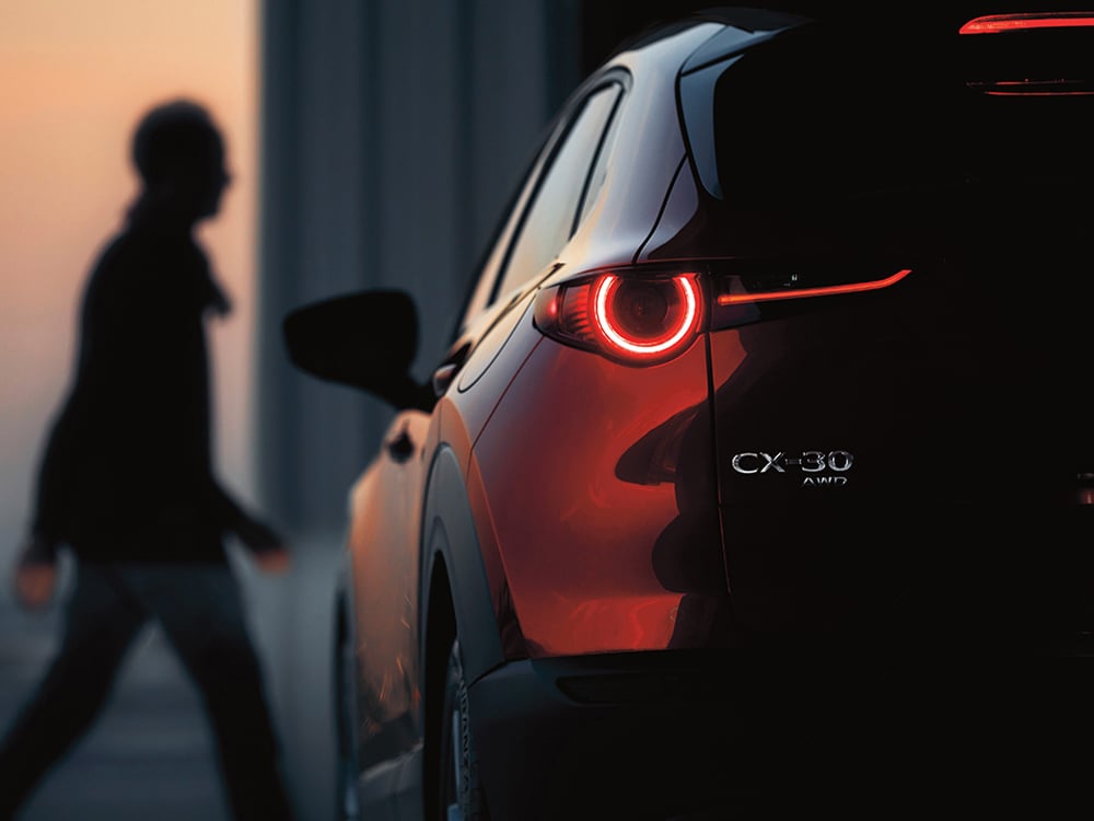 Gros plan de l’arrière côté conducteur d’un CX-30 rouge vibrant cristal métallisé garé au crépuscule, l’accent est mis sur les emblèmes CX-30 et AWD. Le feu arrière est allumé. En arrière-plan, on voit la silhouette floue d’un homme qui marche.