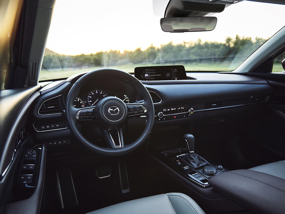 Vue du siège conducteur d’un CX-30 au crépuscule, mettant l’accent sur le volant, le tableau de bord, la console et les commandes des portes.