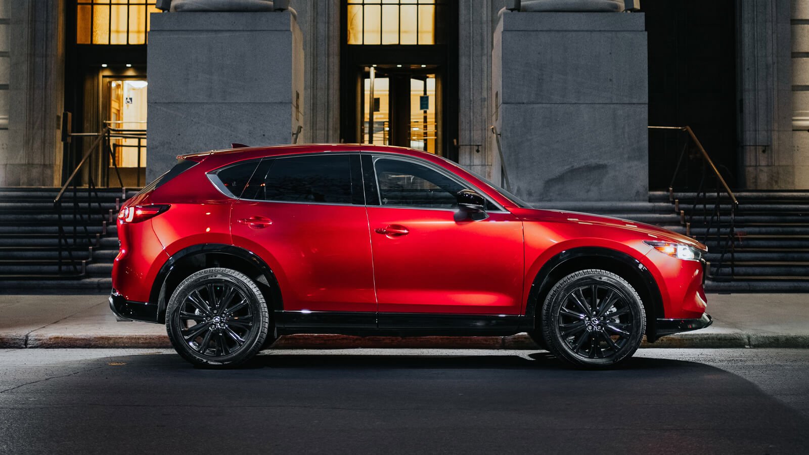 Soul Red Crystal Metallic Mazda CX-5 parked in evening in front of stately historical building, windows and entrance lit warmly.