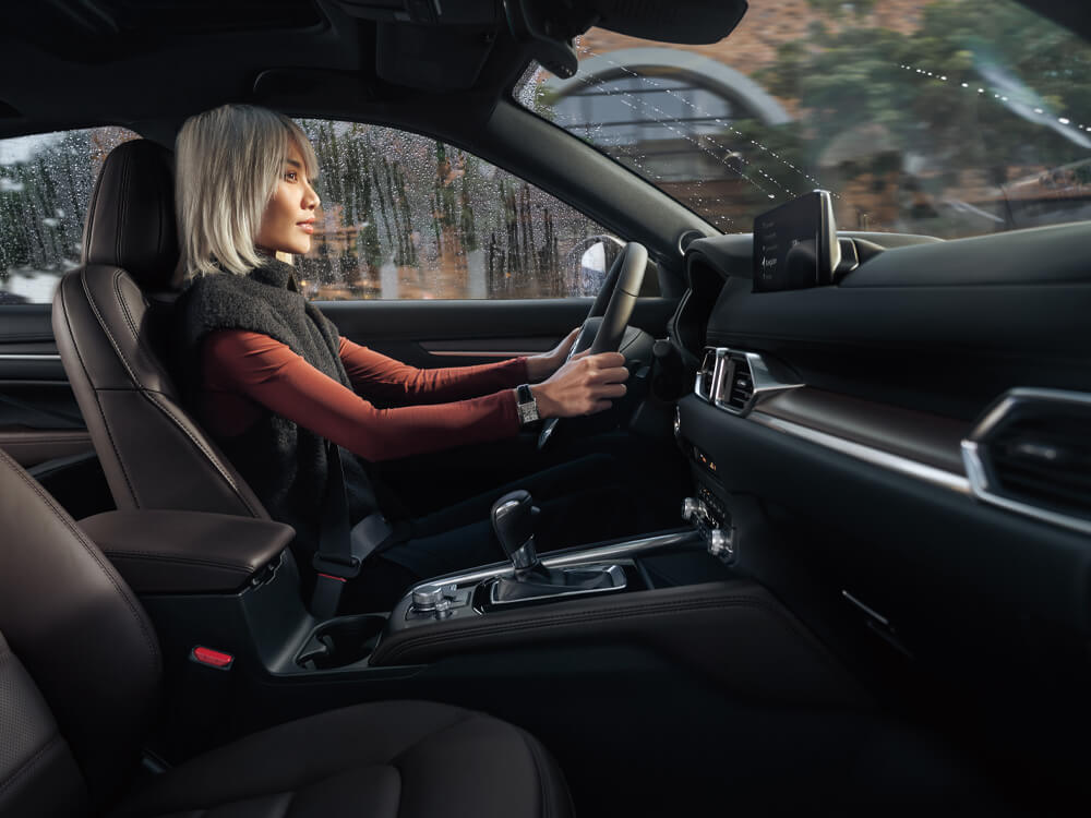 From passenger side, looking across to stylish woman driving CX-5 with Cocoa leather interior.