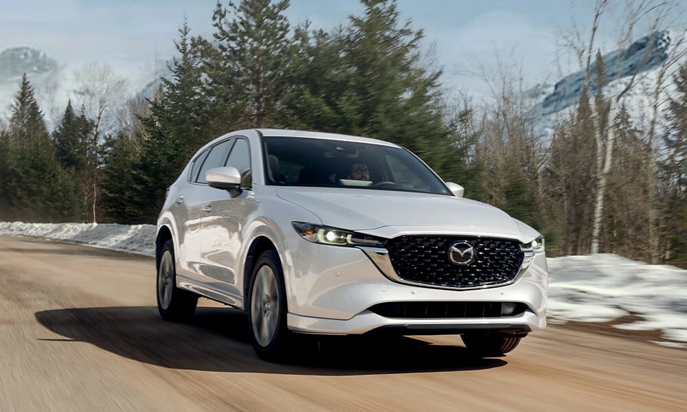 Vue ¾ d’un Mazda CX-5 blanc rhodium métallisé roulant sur une route de pavé avec un arrière-plan enneigé flou.