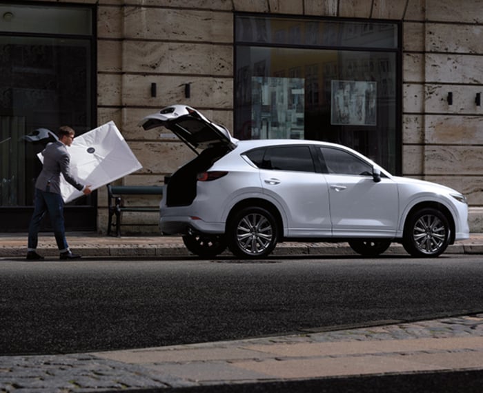 Man carries white frame to Rhodium White Metallic Mazda CX-5 parked on city street with liftgate open. 