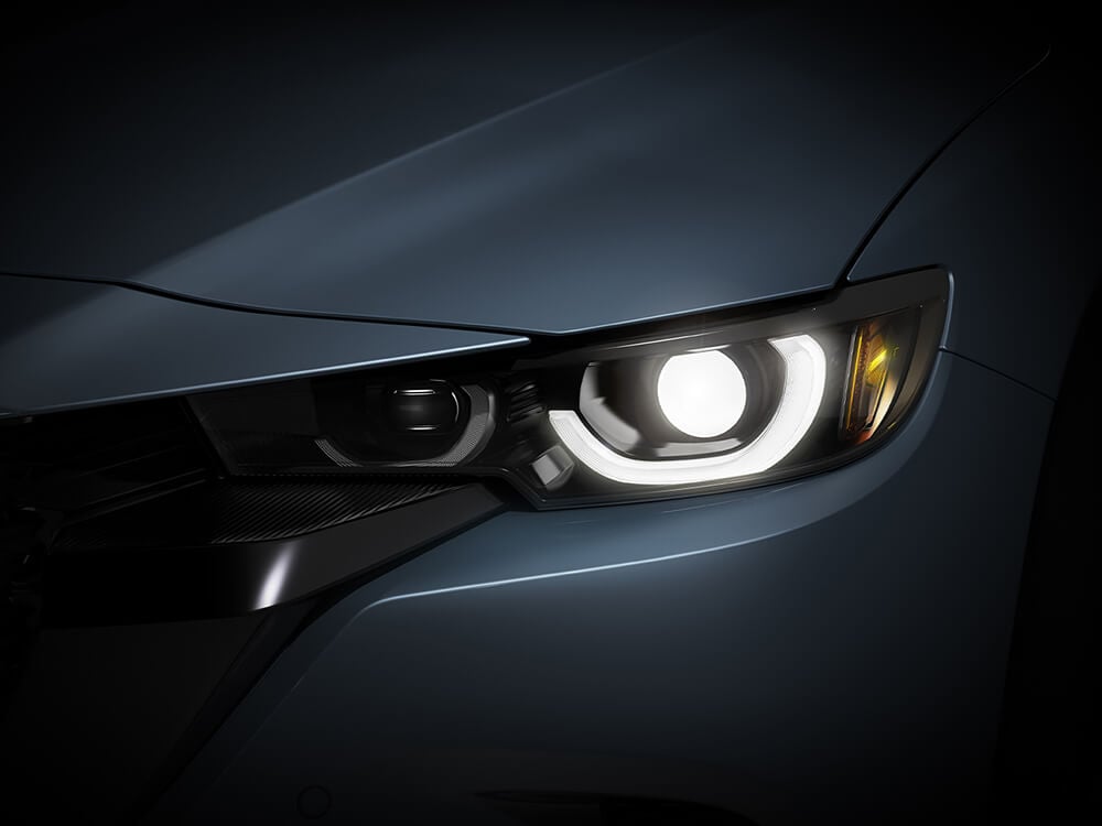 In low light, extreme close up of Mazda CX-50 lit headlamp and surrounding panels and contours.