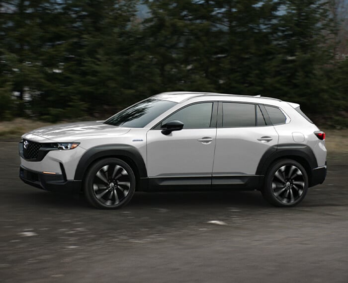 Wind Chill Pearl Mazda CX-50 Hybrid driving in profile close up, trees in background.