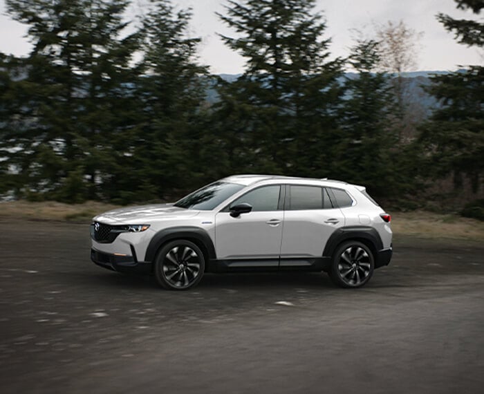 Gros plan de profil d’un Mazda CX-50 hybride vent polaire nacré circulant sur une route avec des arbres en arrière-plan.