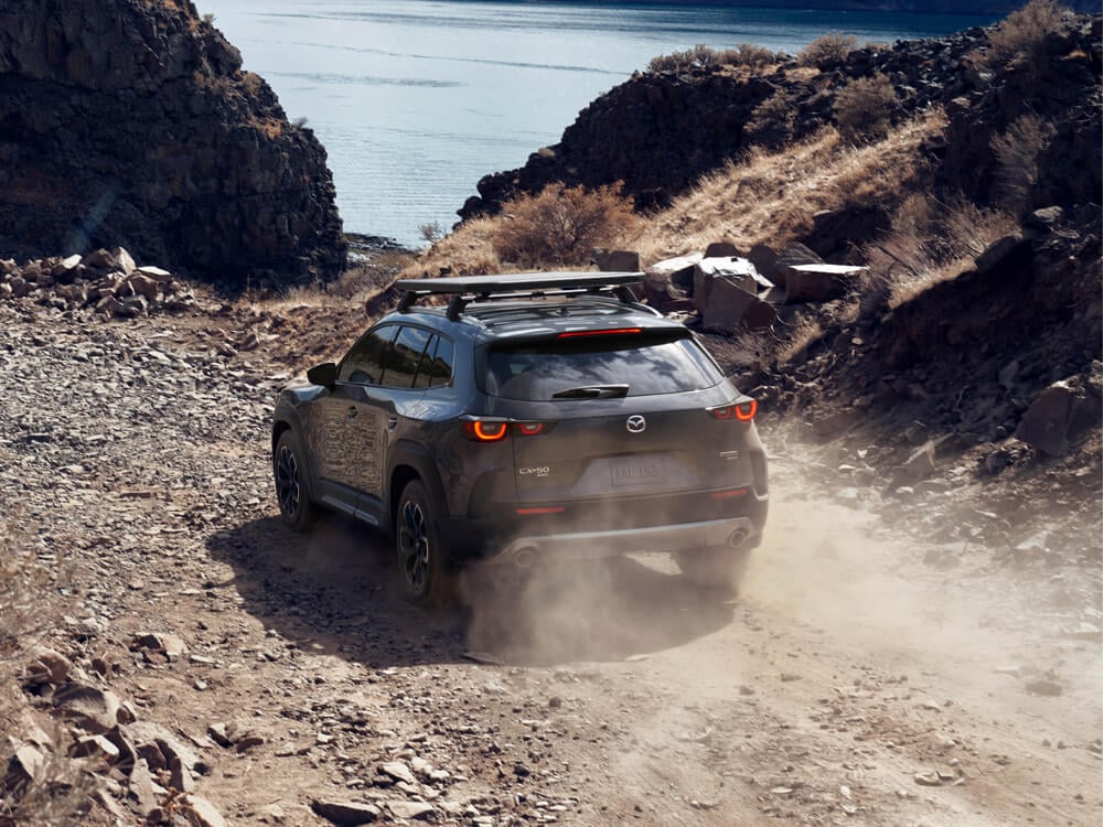 Rear of the Polymetal Grey Metallic Meridian trim CX-50 kicking up dust driving on dirt path away from camera. 