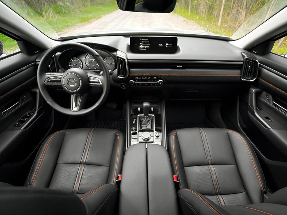 From high above front row seats, looking down at the seats, steering wheel, dash. The focus is orange stitching on the black leather interior. 