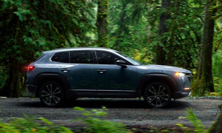 Ingot Blue Metallic CX-50 in profile, driving on gravel path in the forest. 