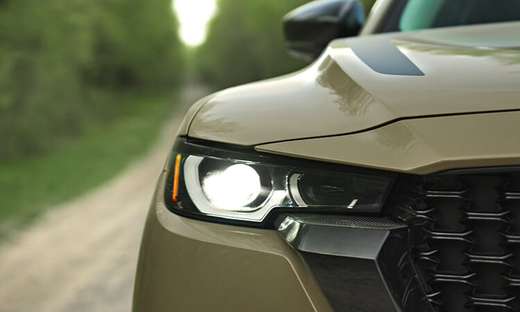 Close up of headlight of Zircon Sand Metallic CX-50 driving toward camera on dirt/gravel road.