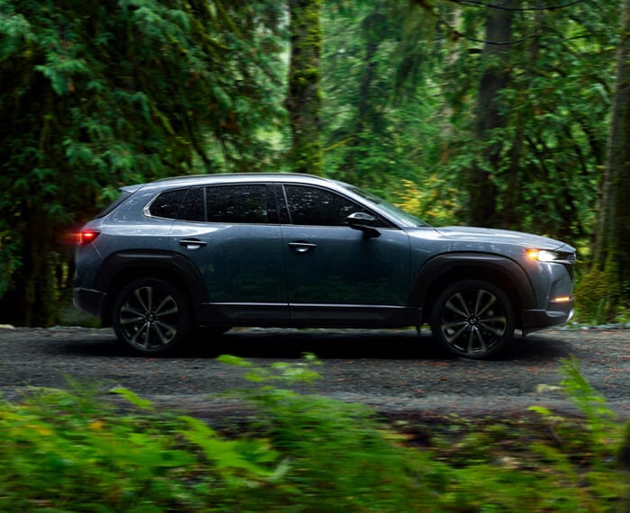 Ingot Blue Metallic CX-50 in profile, driving on gravel path in the forest. 