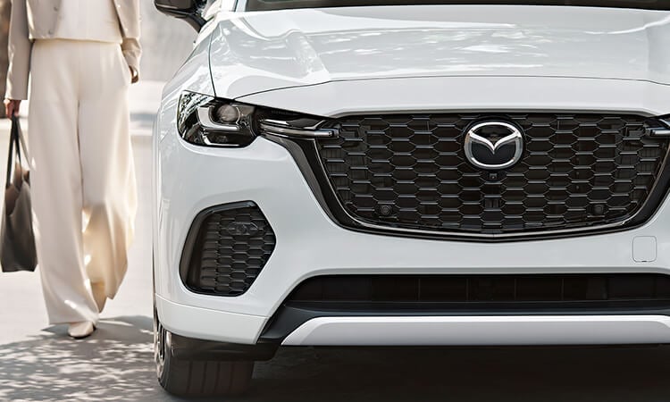 Mid-close up of the front grille and Mazda emblem on a Rhodium White Metallic CX-70, a woman walks alongside the passenger’s side toward the door.