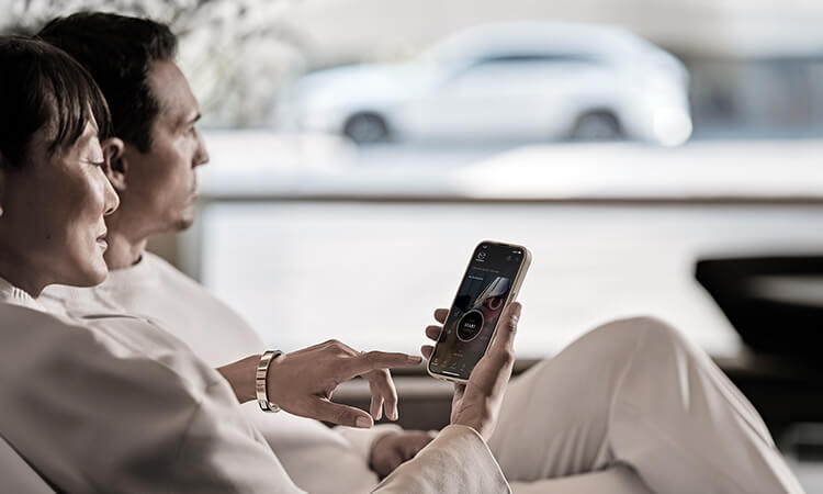A couple sits together in their home, one on her phone with the MyMazda app open. A Rhodium White Metallic CX-70 parked in the background.