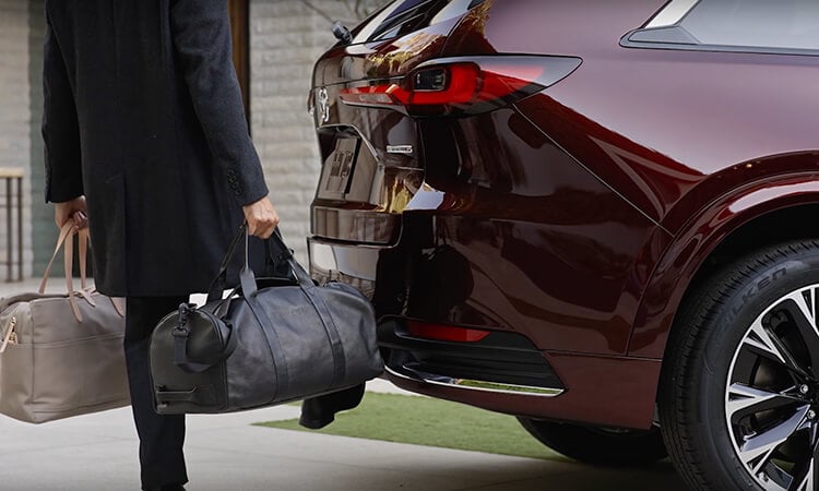 Un homme aux mains pleines utilise son pied pour activer le hayon électrique mains libres d’un CX-90 rouge artisan.