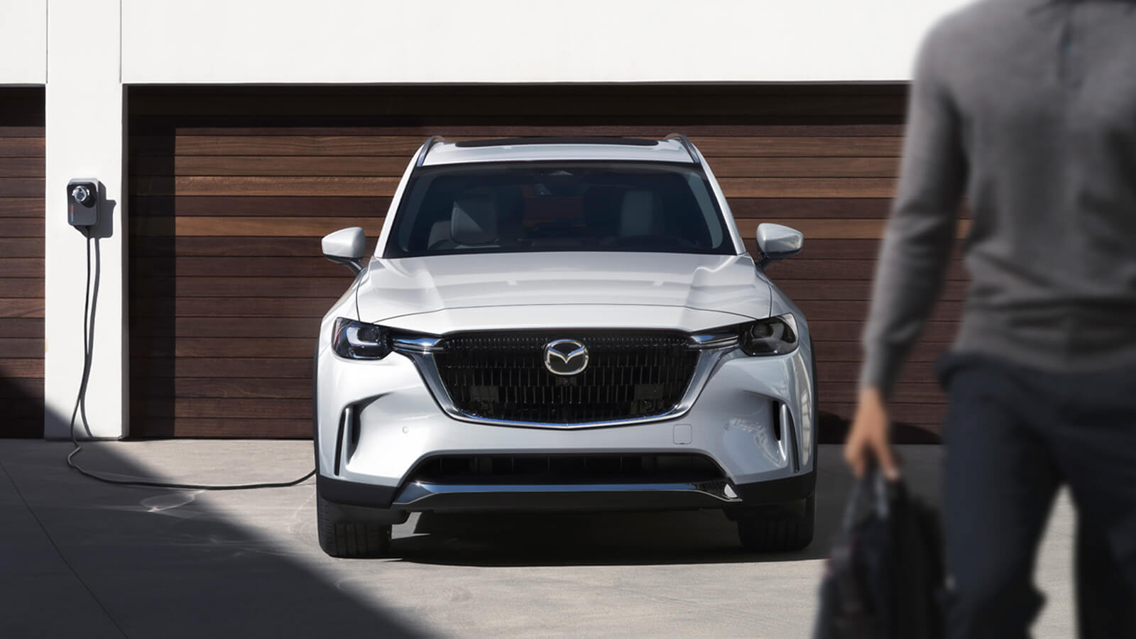 Plugged in and charging Arctic White CX-90 PHEV parked in front of double garage with wood slat garage doors.