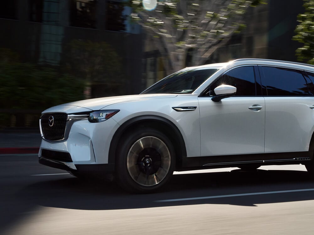 Low front profile shot of Artic white CX-90 driving on street with stately buildings and trees blurred in background. 