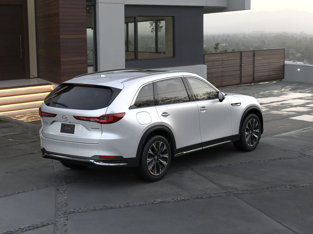 Back ¾ above shot of Arctic White CX-90 parked at an angle in front of stylish modern home. 