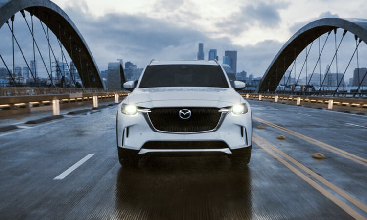 Straight on shot of Arctic White CX-90 driving on a stylish bridge, city skyline in background. 