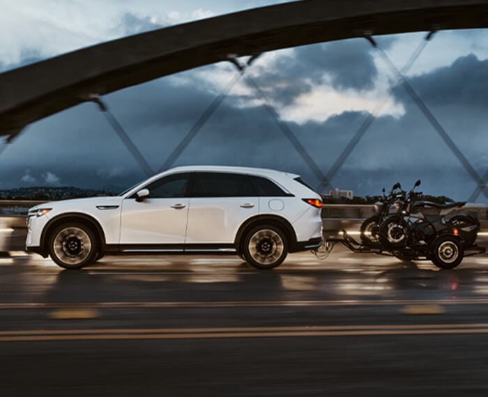 Arctic White CX-90 PHEV driving over a bridge at dusk, towing a small trailer with 2 motorcycles.  