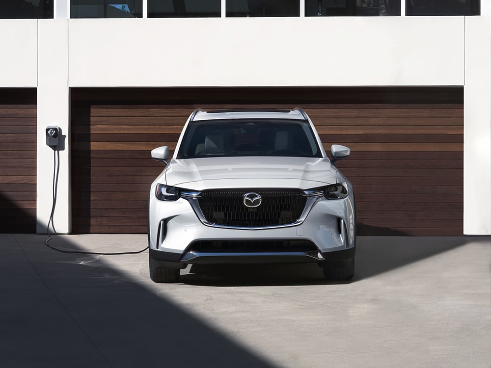 White CX-90 PHEV charging in a home driveway with a wooden slay garage door behind.