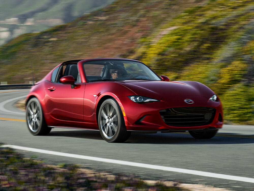 A red MX-5 RF drives down a winding road with scenery blurred in the background.