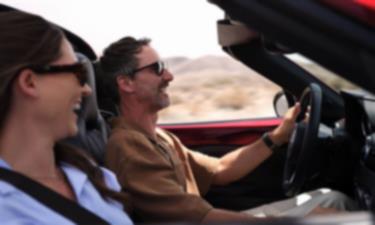 Two smiling people in sunglasses drive down a road in a red MX-5 RF.