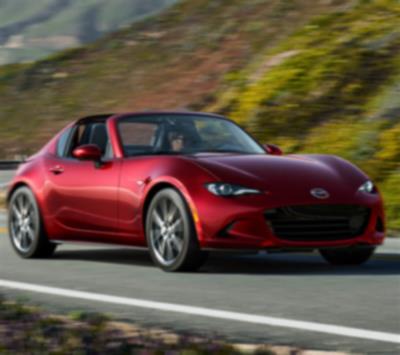 A red MX-5 RF drives down a winding road surrounded by hilly terrain. 