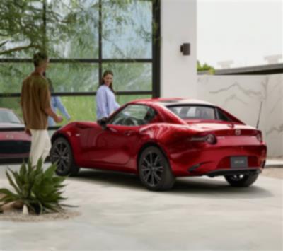 Two people look at a red MX-5 RF parked in front of a reflective window.