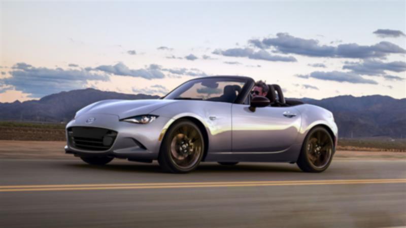 A silver MX-5 Soft Top with its top down drives down an open road with mountains visible.